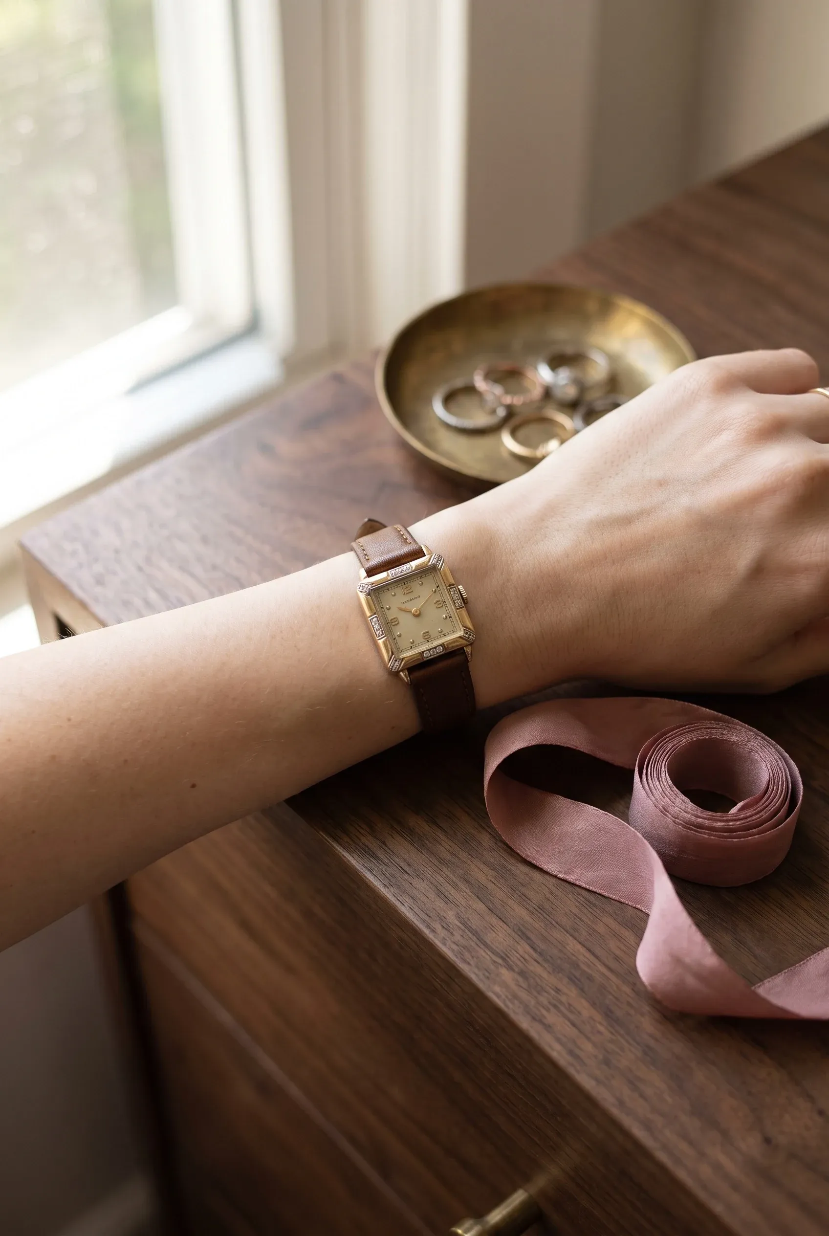 Petite wrist with a square stone-set watch beside a ring tray and ribbon on a dresser.
