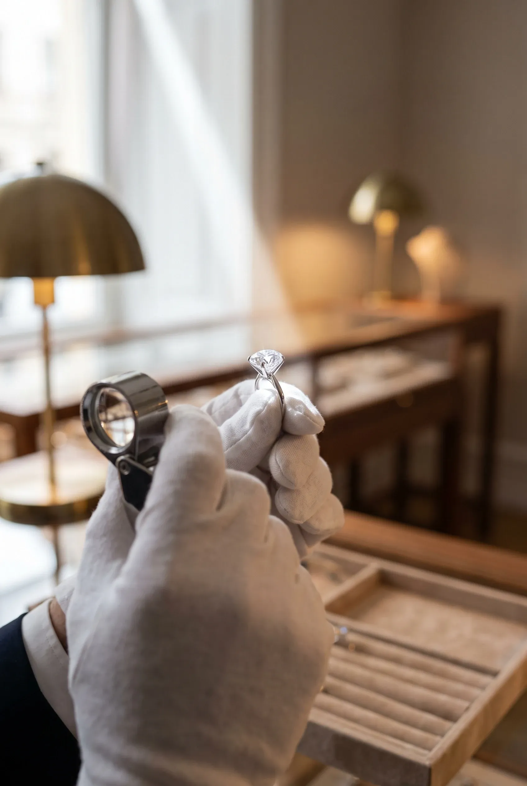 White-gloved jeweler holding a solitaire ring up to window light, a loupe ready in the other hand