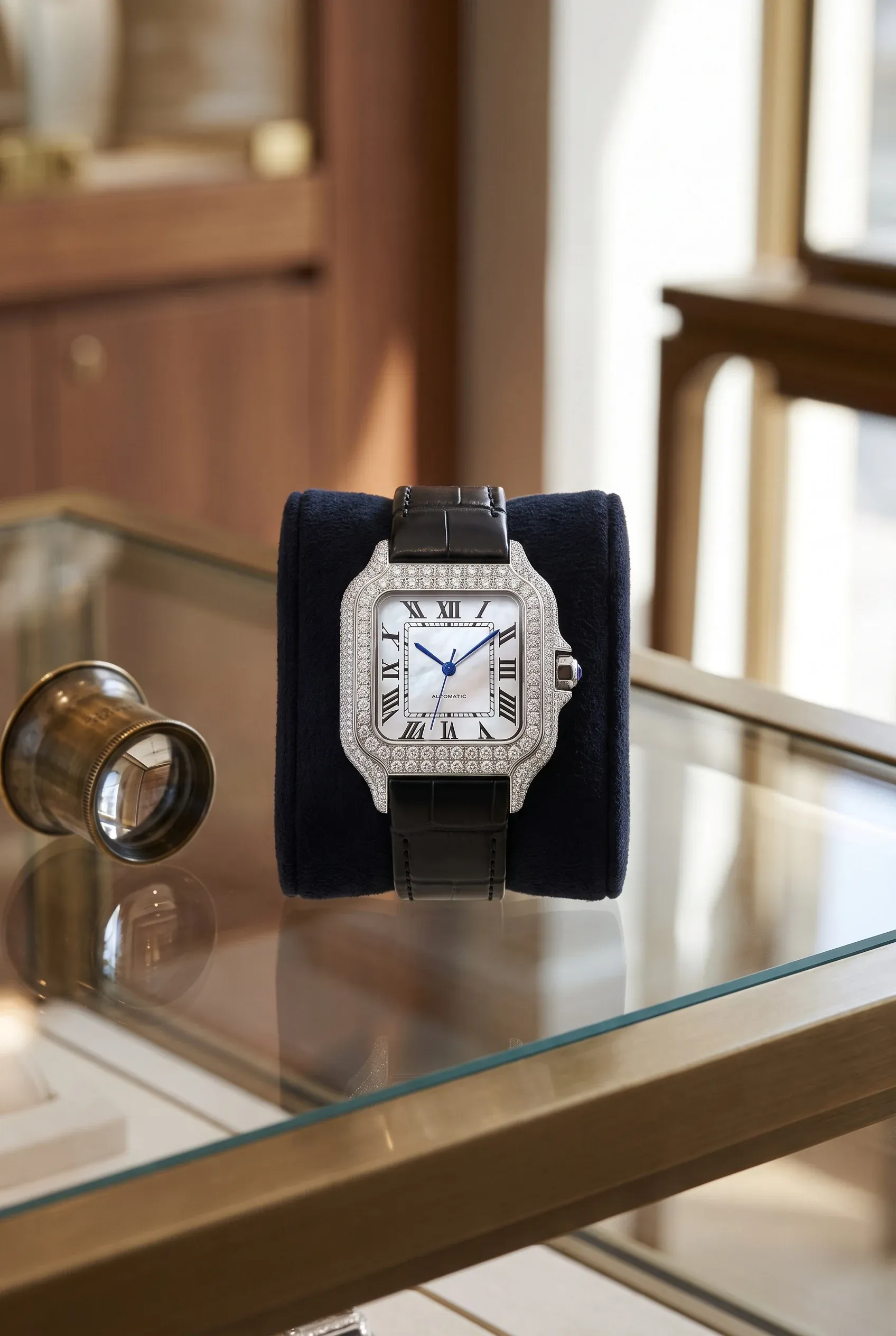 Stone-set square watch with a mother-of-pearl dial on a navy velvet cushion beside a brass loupe on a glass counter
